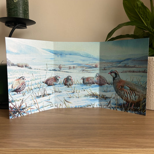 Trifold greetings card featuring a painting of partridges on the downs displayed on a wooden surface.