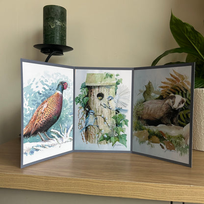 Three-panel nature-themed triptych greetings card on a wooden surface with a lamp and plant in the background.