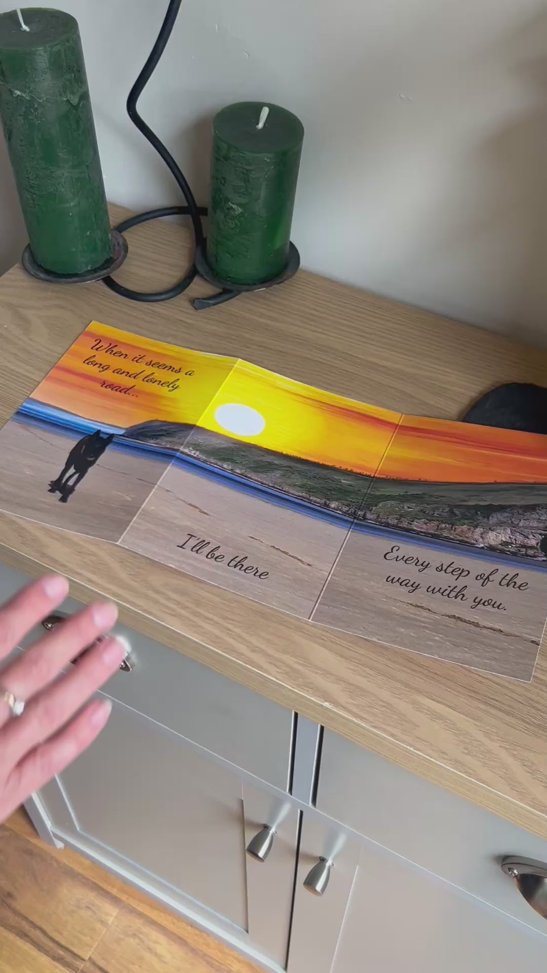 The video shows a demonstration of the Bare Cards Trifold Dog Photo Sentiment Card. The trifold image opens up to show a stunning sunset across a deserted beach and a dog trotting towards the camera. The caption reads "When it seems a long and lonely road... I'll be there every step of the way". The video then shows how to fold and seal this unique card to form the built-in envelope depicting sunset over a field of corn.
