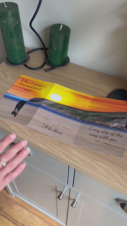 The video shows a demonstration of the Bare Cards Trifold Dog Photo Sentiment Card. The trifold image opens up to show a stunning sunset across a deserted beach and a dog trotting towards the camera. The caption reads "When it seems a long and lonely road... I'll be there every step of the way". The video then shows how to fold and seal this unique card to form the built-in envelope depicting sunset over a field of corn.