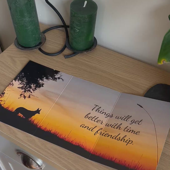 The video shows a demonstration of the Bare Cards Trifold Dog Photo Sentiment Card. The trifold image opens up to show a stunning sunset and silhouette of a dog under a tree and the caption "Things will get better with time and friendship". The video then shows how to fold and seal this unique card to form the built-in envelope depicting sunset over a field of corn.